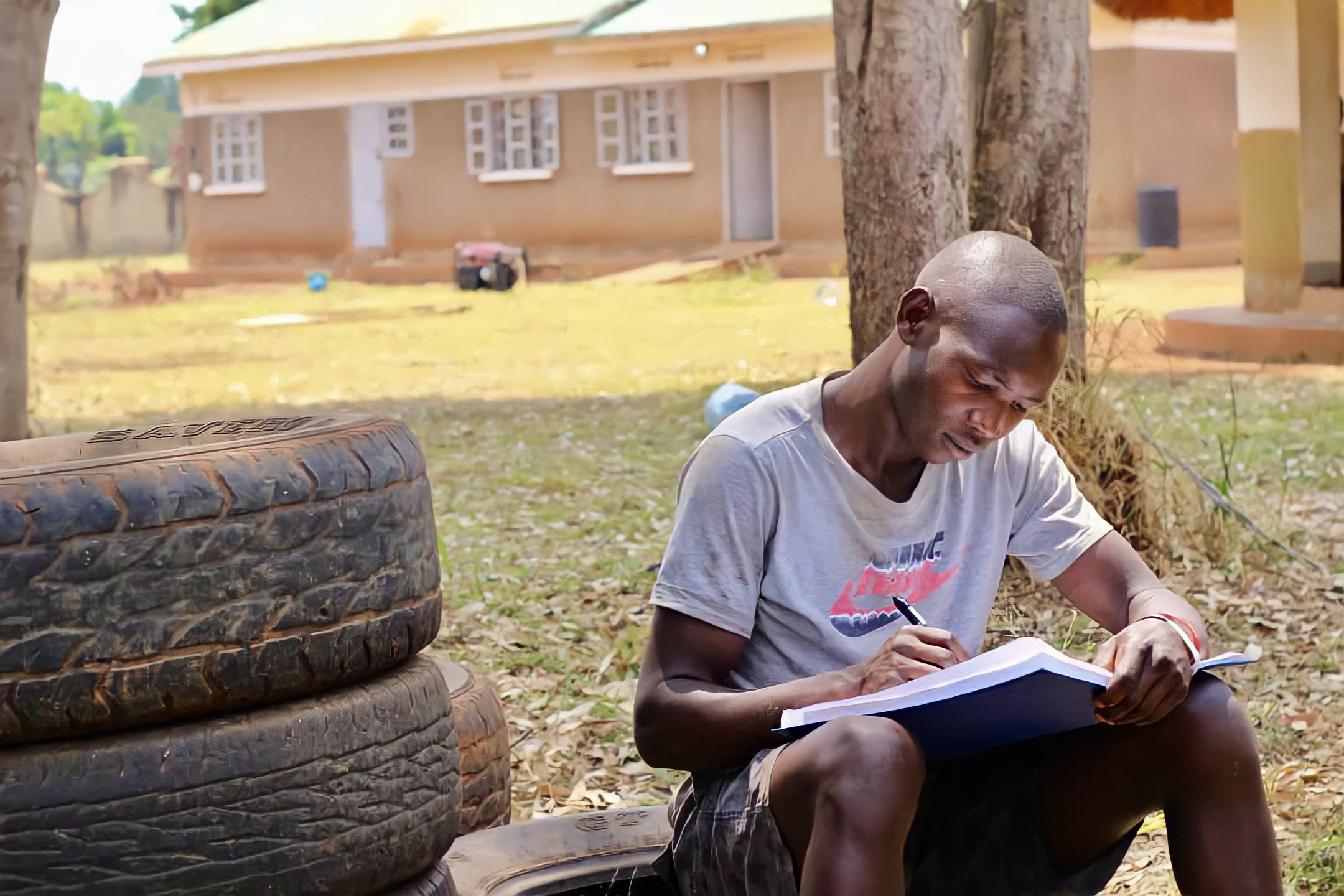 A young man sits outside on an old tire next to a pile of three more tires. He is writing in a notebook.