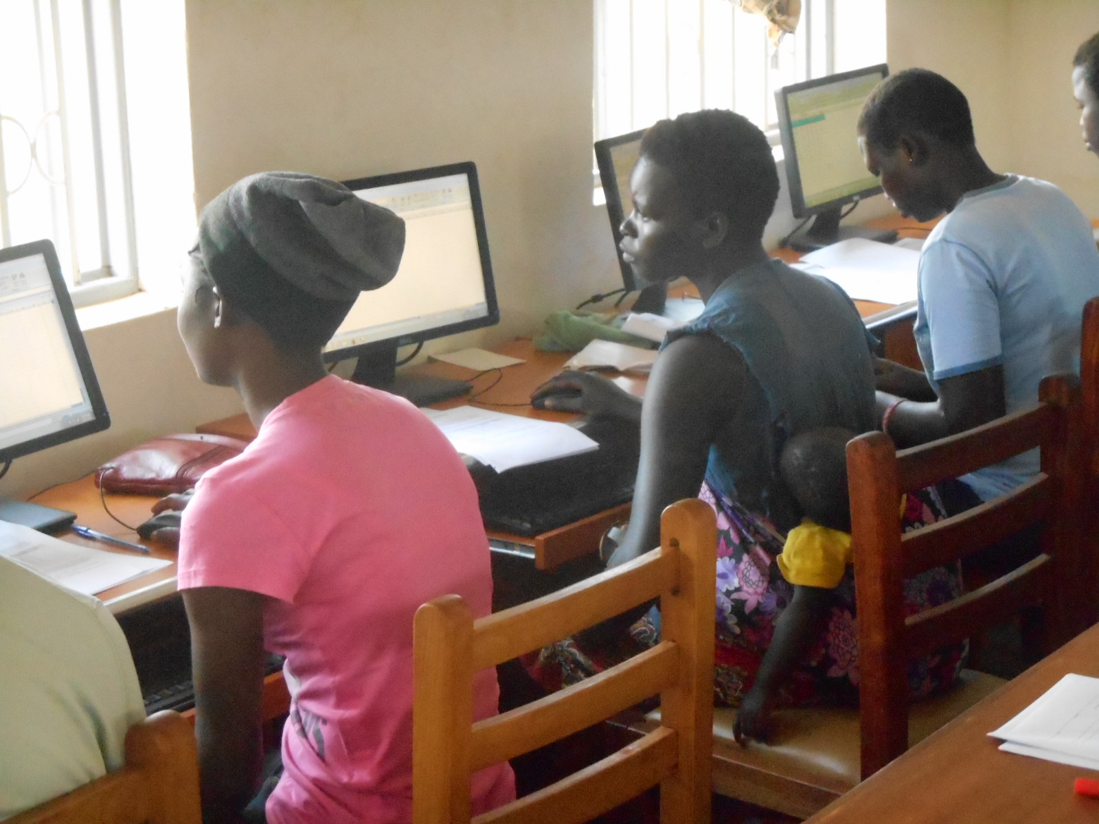 Three young people sitting at a row of computers. The young woman in the middle has a sleeping baby secured to her back.