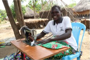 Almar sits behind her sewing machine, smiling, as she feeds fabric under the stitching foot.