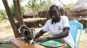 Almar sits behind her sewing machine, smiling, as she feeds fabric under the stitching foot.